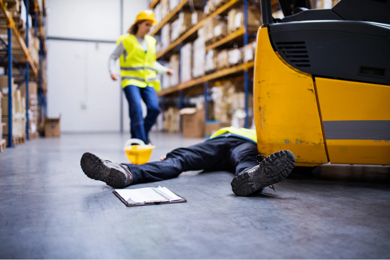 A worker lies on the floor next to a forklift in a warehouse as another worker rushes towards them.