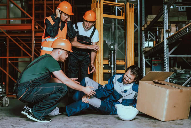 Workers assist a colleague who has fallen beside a forklift in a warehouse.