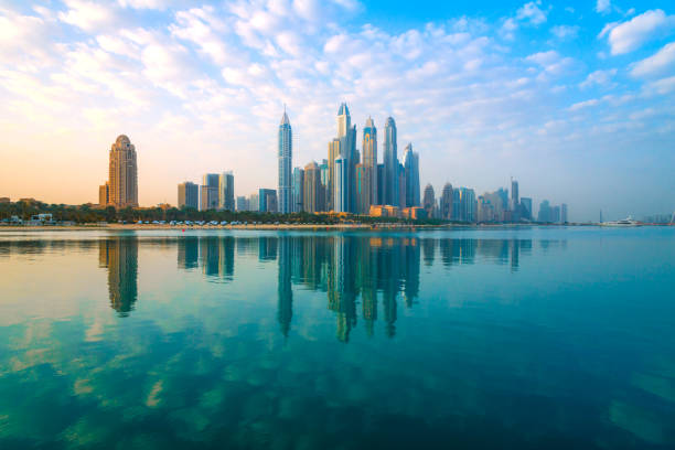 Skyline of Dubai with tall buildings reflected in tranquil water under a partly cloudy sky.