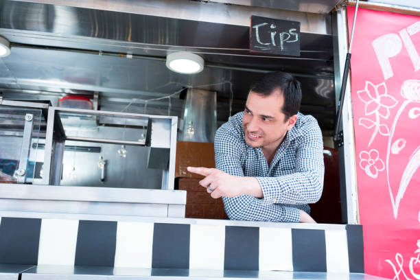 A man in a checkered shirt leans out of a food truck window and points outside.